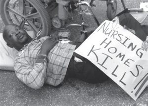 man chained to his wheel chair laying on the ground with a sign that says Nursing Homes Kill.