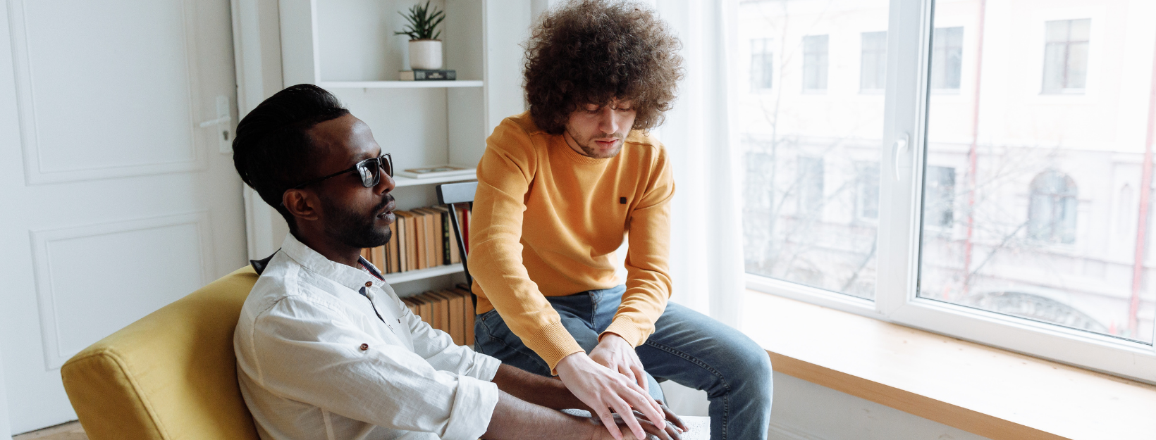 white man with curly hair and orange shirt is teaching a blind black man wearing a dress shirt how to read Braille.. The teacher's hands are guiding the student's as he tries to read a book