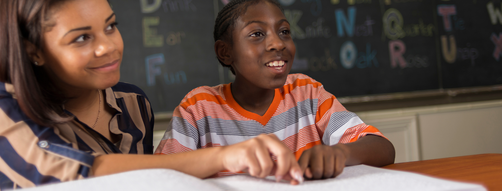 In a classroom, a young blind black male student wearing orange striped shirt who is learning to read Braille from his female teacher who is wearing a blue blouse with brown stripes