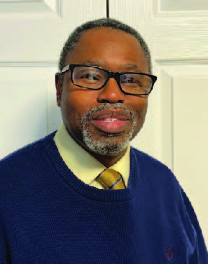 Portrait of a man with glasses, dark gray hair and beard. He is smiling and wearing button down shirt and tie under a sweater.