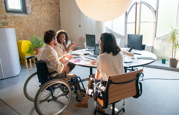 Three people sitting at a table in a bright office setting. Two are sitting in chairs and one in a wheelchair with a laptop in front of him.