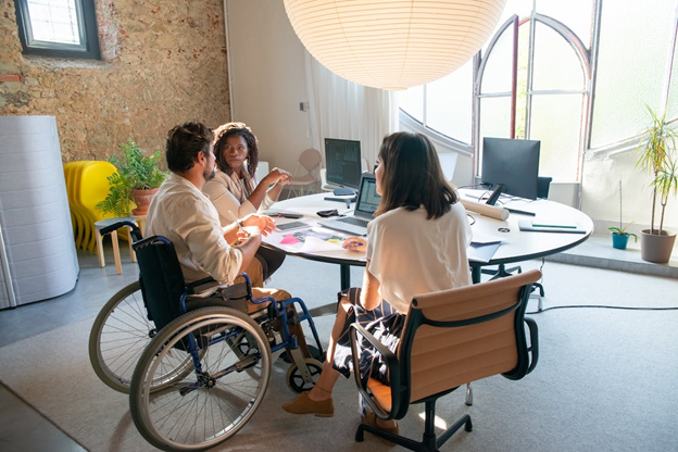 Three people sitting at a table in a bright office setting. Two are sitting in chairs and one in a wheelchair with a laptop in front of him.