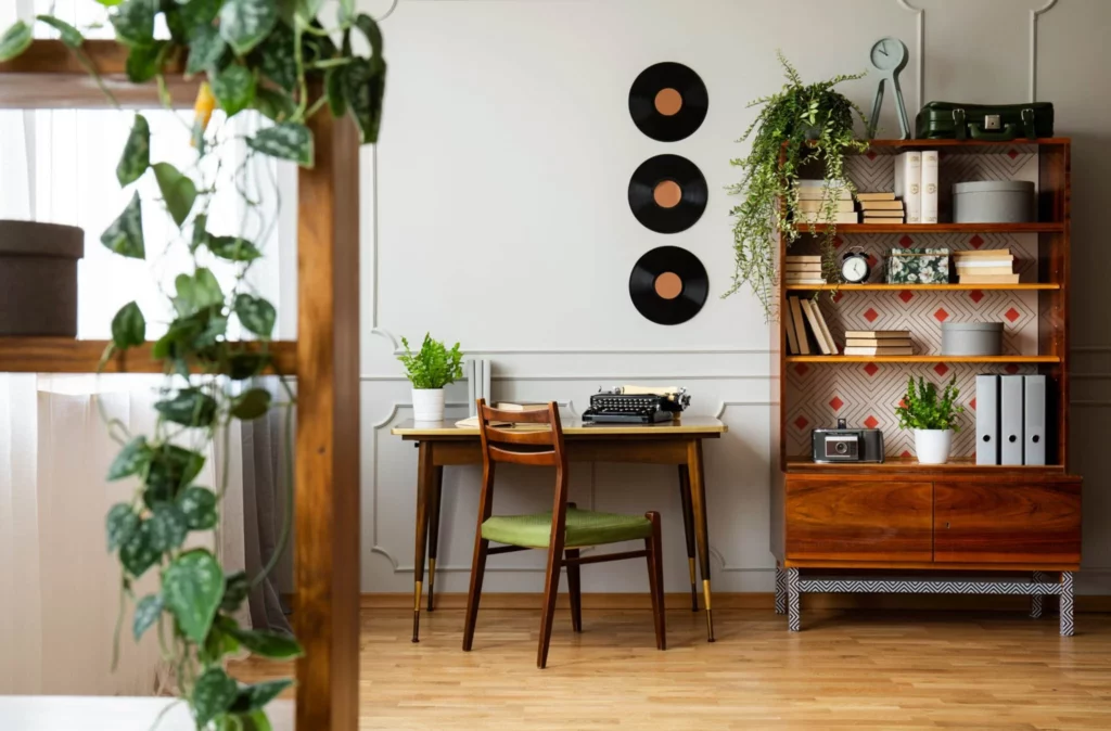 A corner of a room with light coming through the window on the left. In the center of the room there is a wooden desk with a typewriter. There is a bookcase in the foreground and one in the background with books. There are plants on the bookcases and the desk. The walls are white and decorated with records.