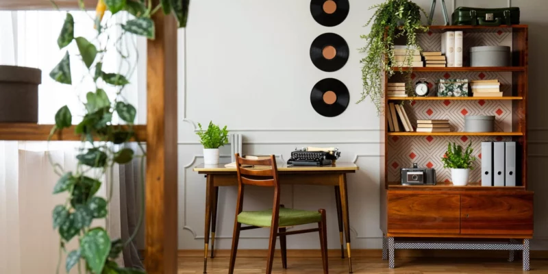 A corner of a room with light coming through the window on the left. In the center of the room there is a wooden desk with a typewriter. There is a bookcase in the foreground and one in the background with books. There are plants on the bookcases and the desk. The walls are white and decorated with records.