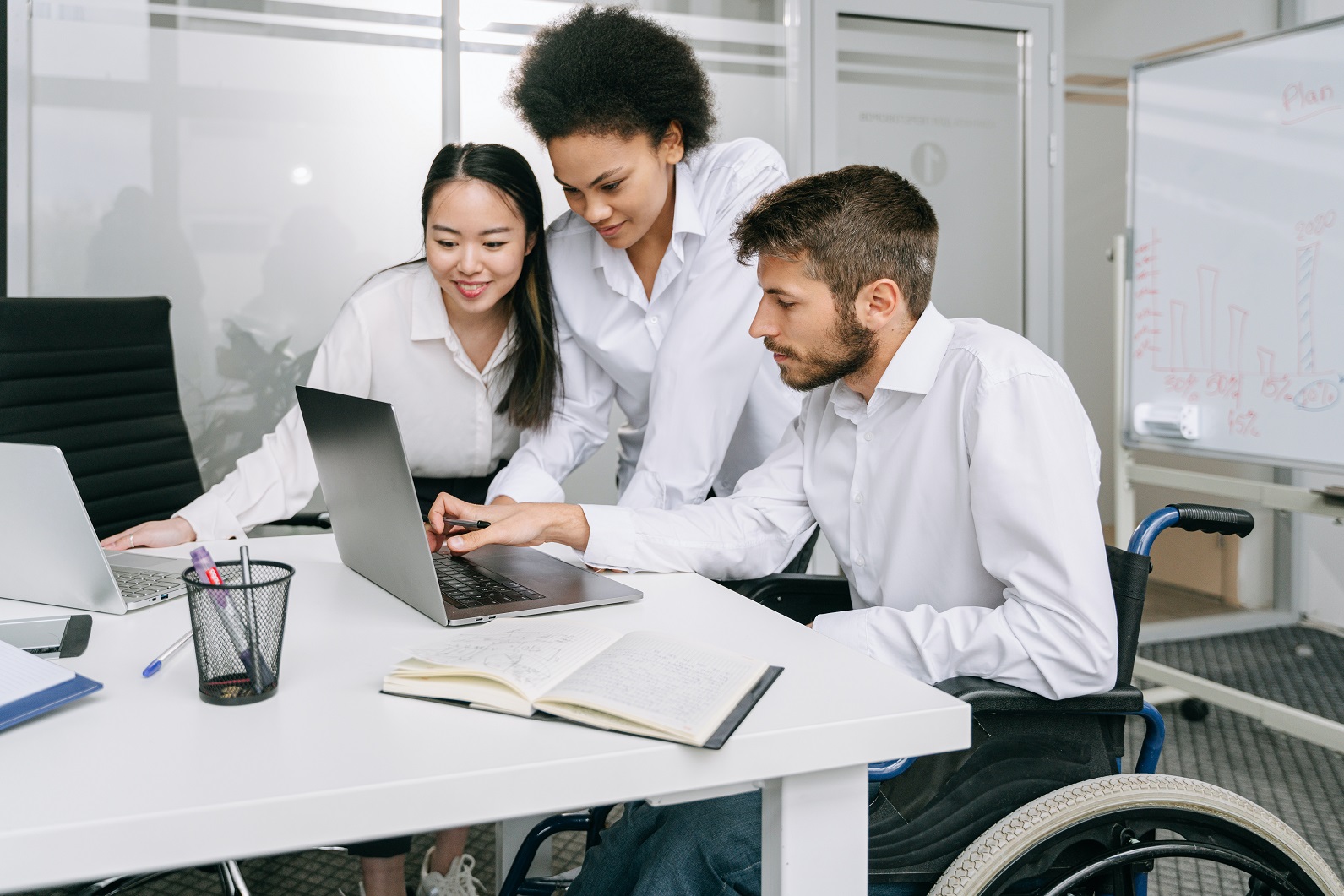 Three women in an office setting. A man sitting in a wheelchair typing on a laptop, and two women standing next to them looking at the screen.