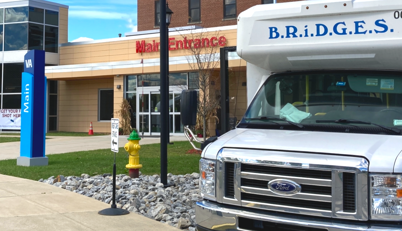 A bus with Bridges written on the front parked in front of a building. Above the door on the building says "Main Entrance."