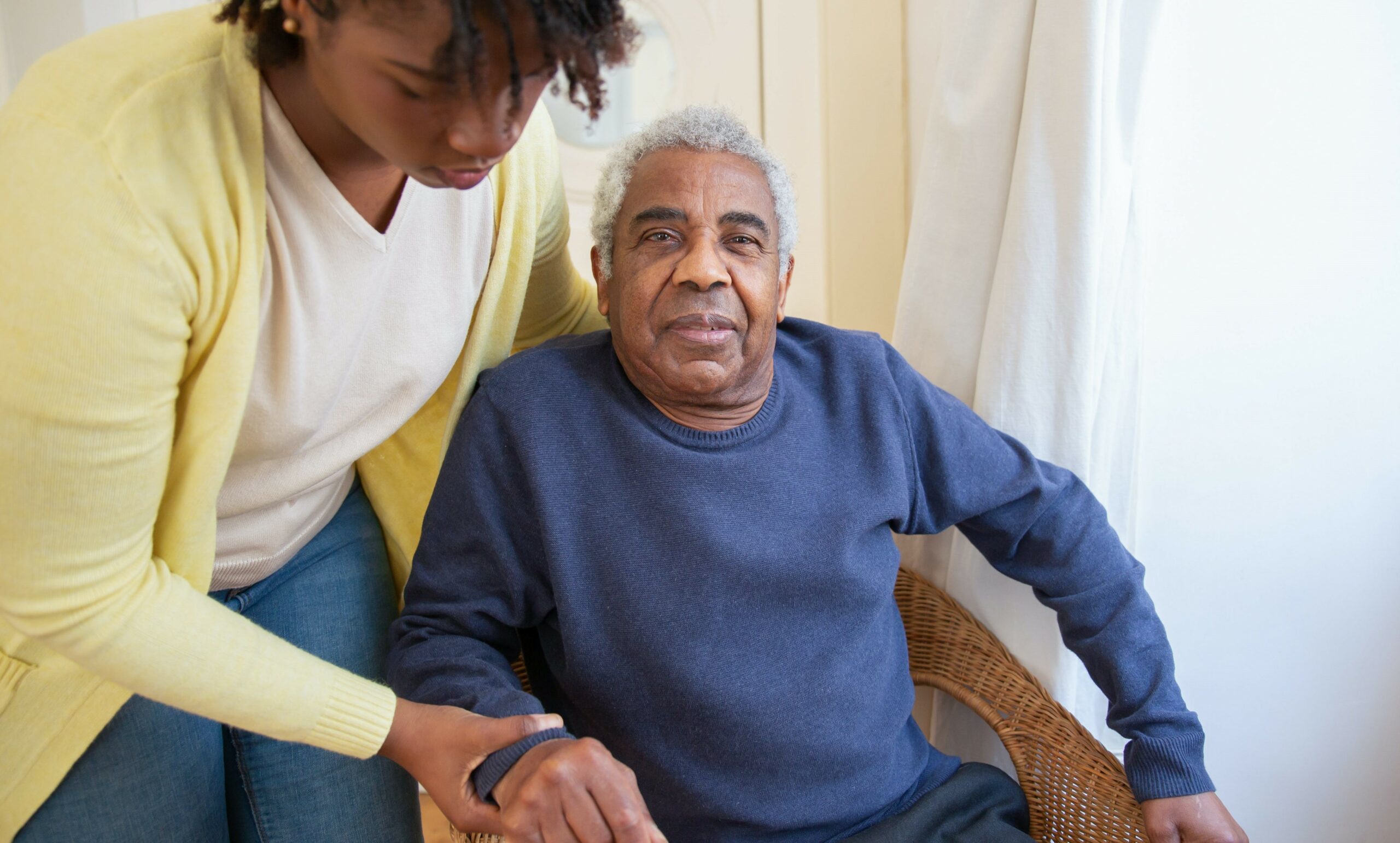 An old man sitting in a chair while a young woman stands next to him holding his arm to assist him.